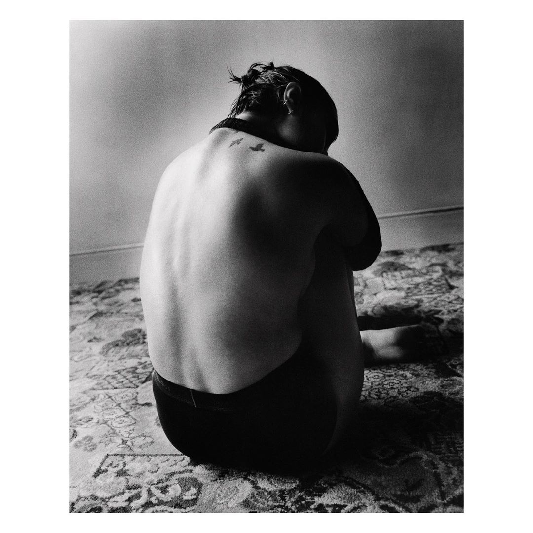 Back view of a woman with a bird tattoo on her back, seated on a patterned carpet in a room with a visible wall.