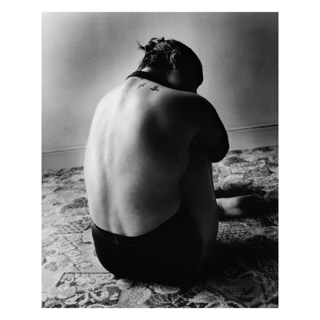 Back view of a woman with a bird tattoo on her back, seated on a patterned carpet in a room with a visible wall.