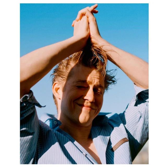 Smiling man in a blue striped shirt poses against a clear sky, holding his hands above his head in a fashion-style portrait.
