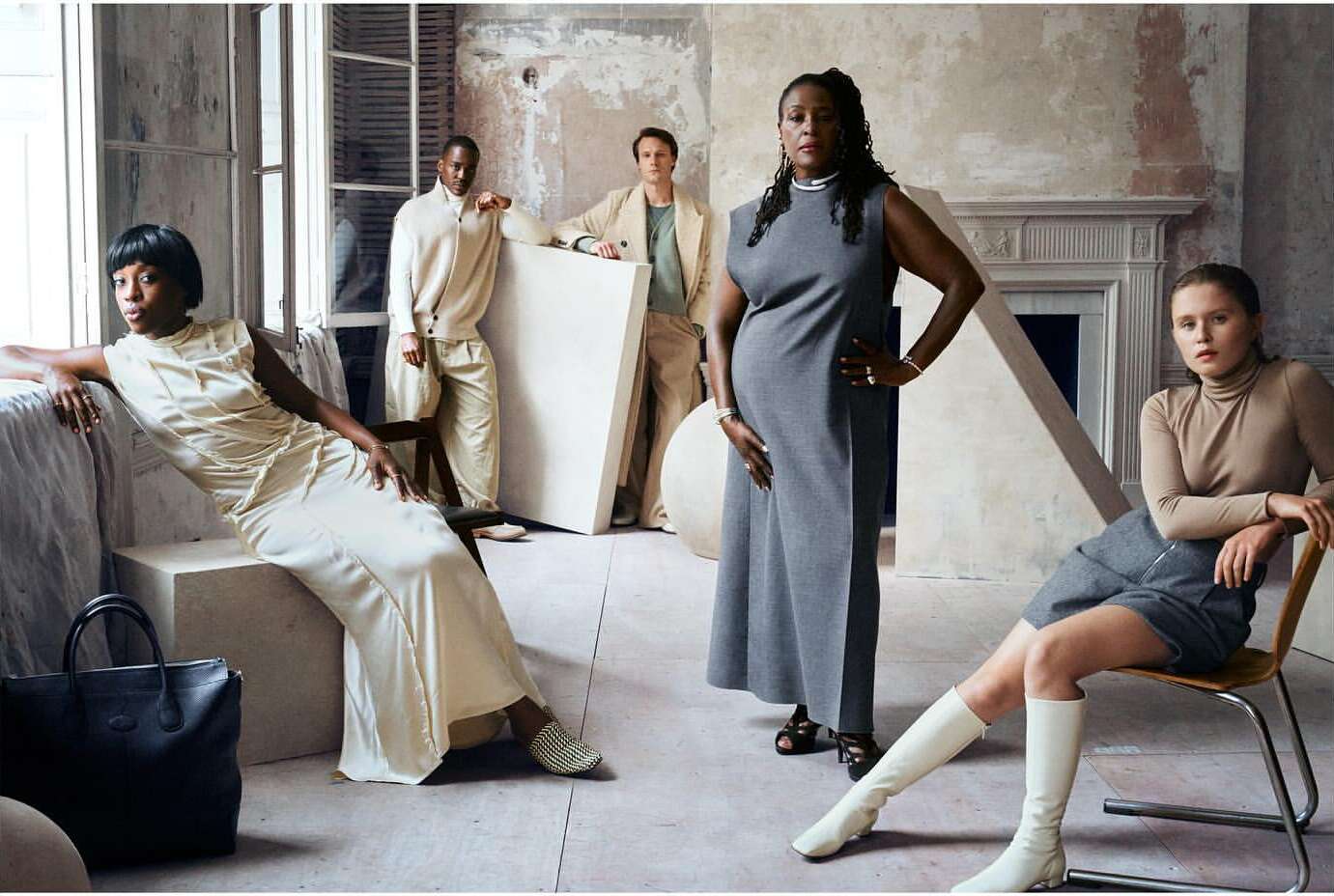 Group of five diverse fashion models in neutral-toned clothing in a bright indoor setting with props and painted backdrop.