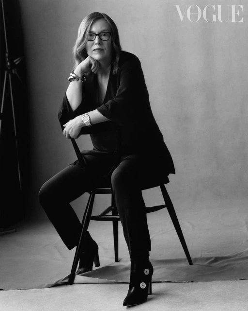 Black and white portrait of a mature woman seated on a chair, wearing glasses and a dark suit, with the Vogue logo visible.