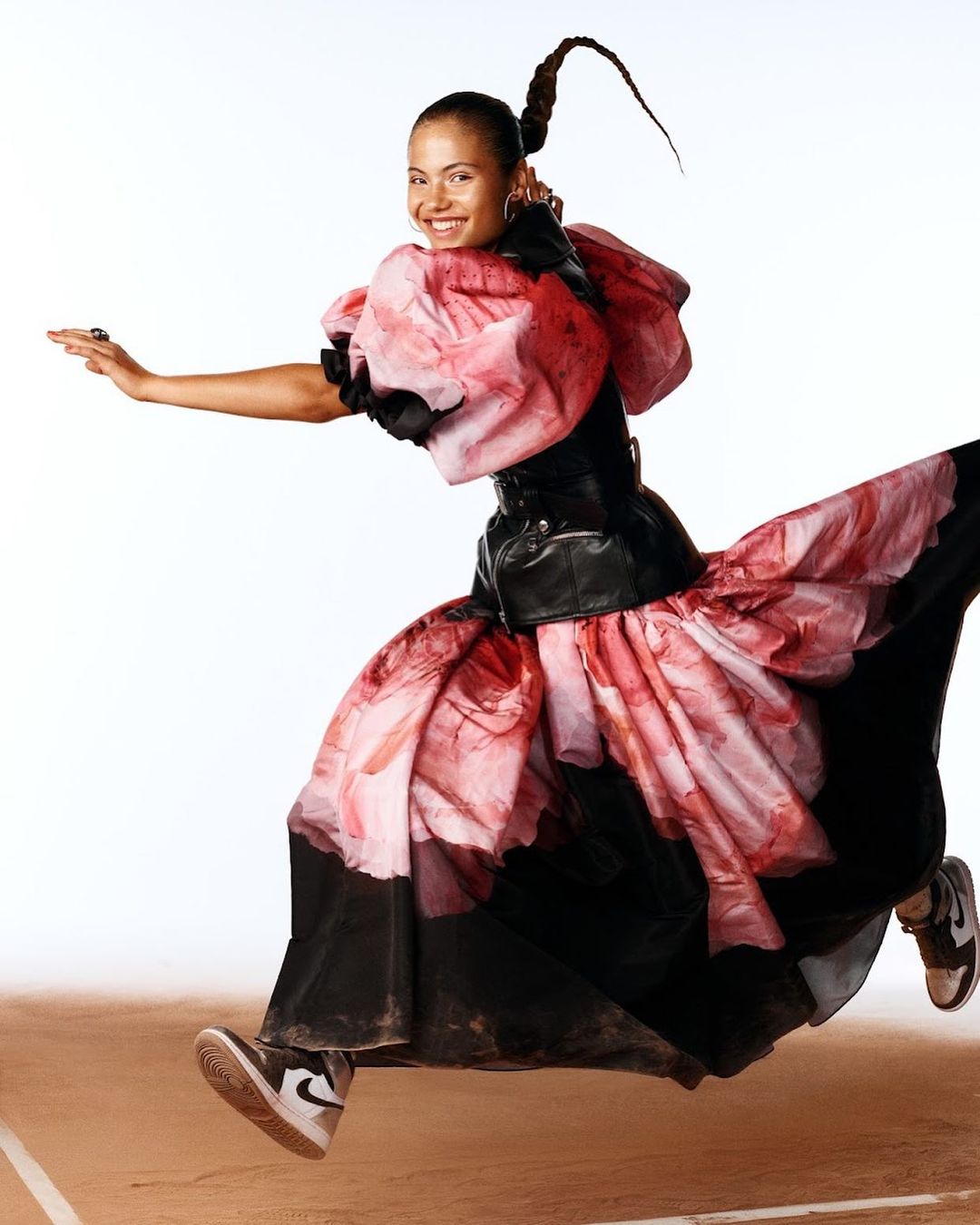 Woman in floral couture gown and sneakers leaping against a gradient background, smiling with arms outstretched in a dynamic pose.
