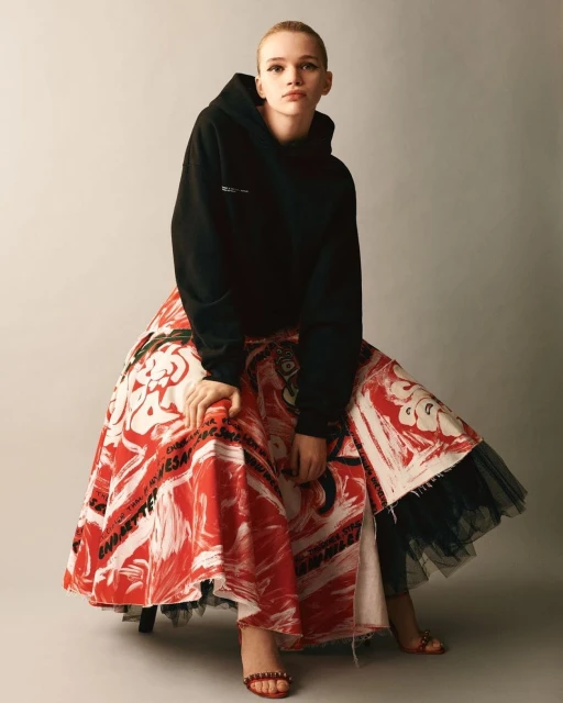 Young woman sits wearing a black hooded sweatshirt and an artistic white-and-red printed skirt, styled for a contemporary fashion shoot.