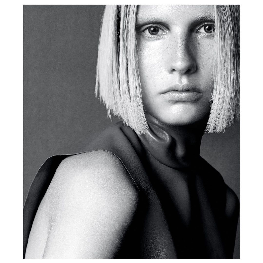 Portrait of a fair-skinned woman with a bob haircut posing in a sleeveless, leather top against a dark grey backdrop in a studio.