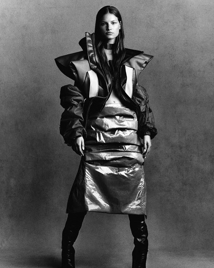 A fashion model poses in a photo studio wearing an avant-garde outfit with knee-high boots, against a gray gradient background.