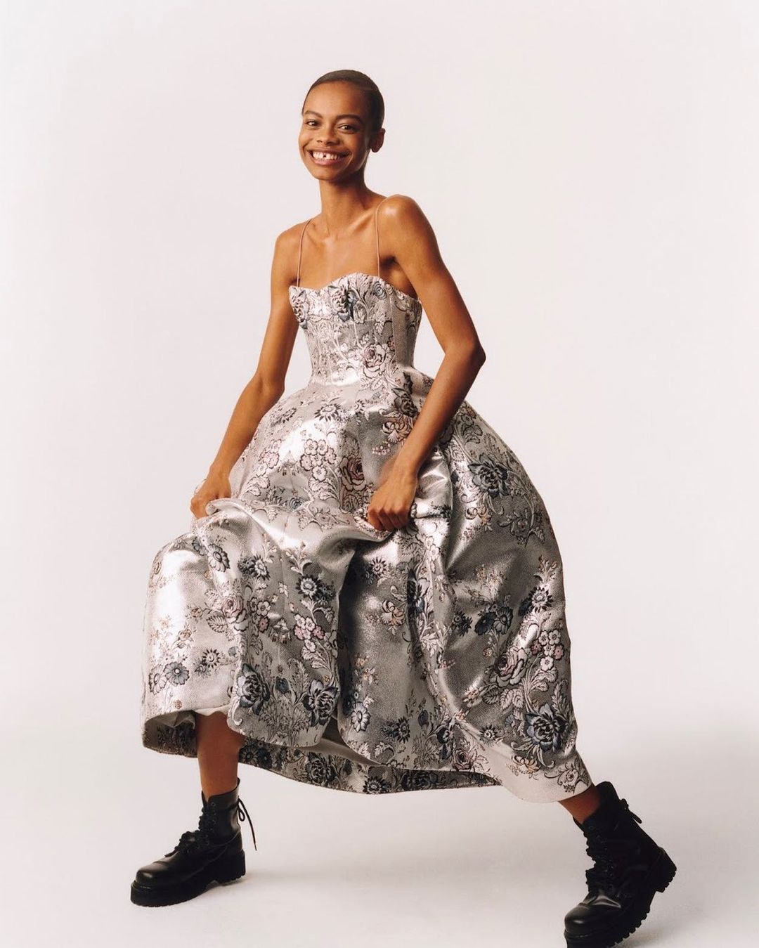 Fashion model poses in a silver floral gown and black platform boots in a studio setting, smiling confidently at the camera.