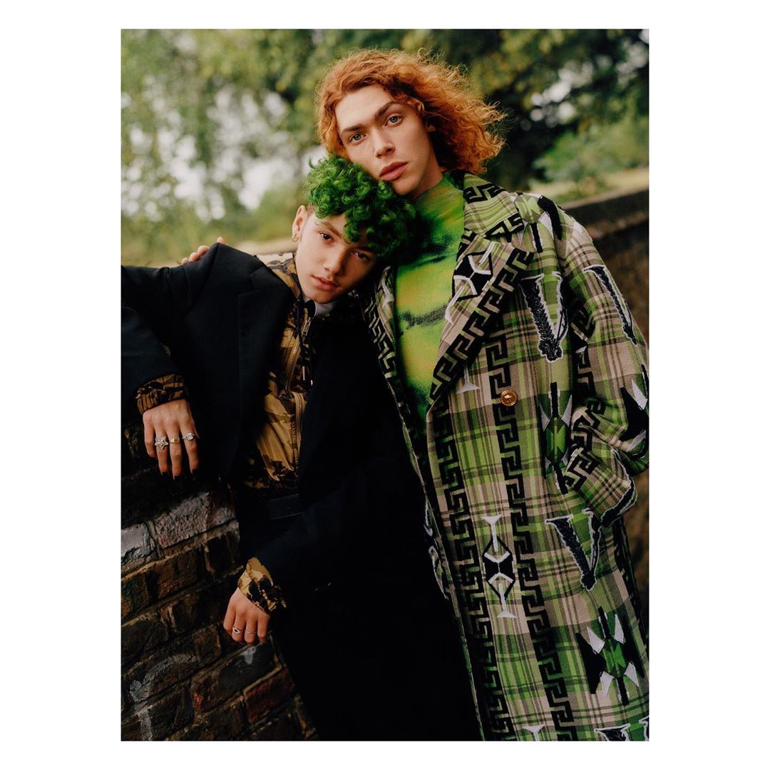 Two young adults, one with green hair and the other with red hair, pose in patterned jackets against a brick wall outdoors.