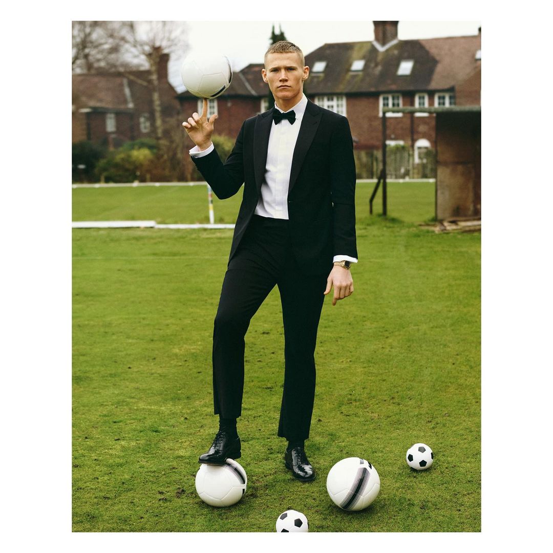 Man in a black suit balances a soccer ball on his finger, standing on a grass field with more soccer balls. Outdoor daytime setting.