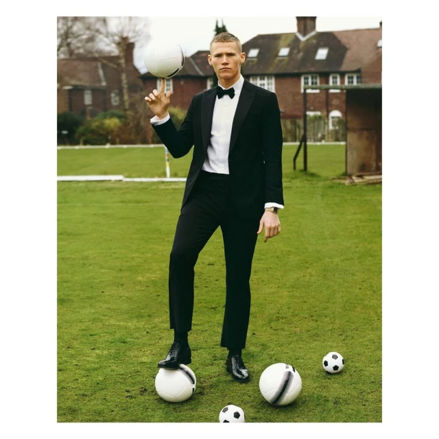 Man in a black suit balances a soccer ball on his finger, standing on a grass field with more soccer balls. Outdoor daytime setting.