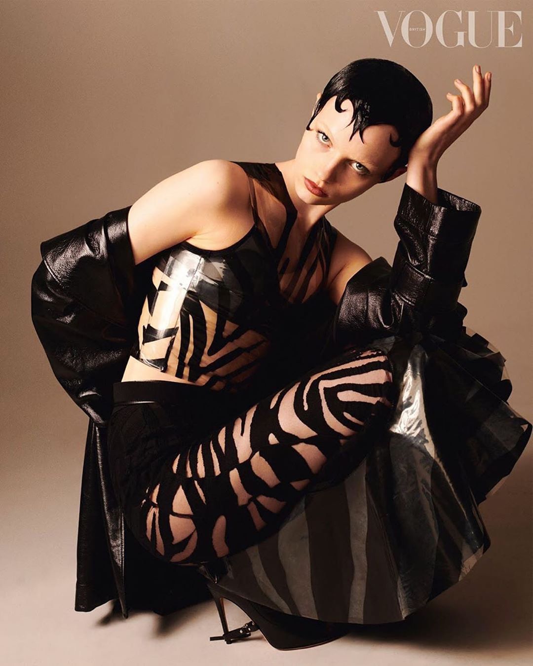 A striking fashion shot featuring a model in a glossy black outfit with zebra print details, crouching indoors with a Vogue logo visible.