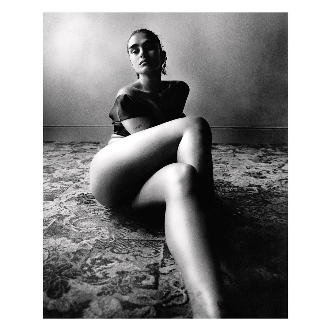 Black and white portrait of a woman sitting on a floral patterned rug, with a neutral wall backdrop, creating a high-contrast, intimate atmosphere.