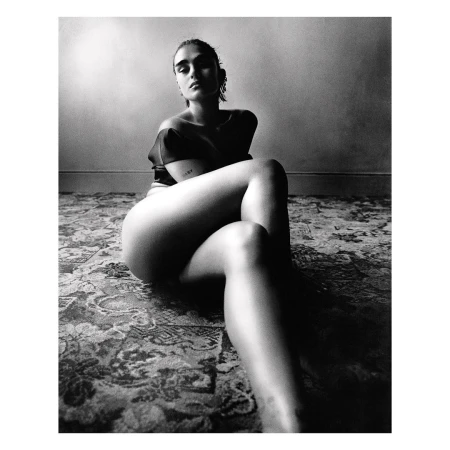 Black and white portrait of a woman sitting on a floral patterned rug, with a neutral wall backdrop, creating a high-contrast, intimate atmosphere.
