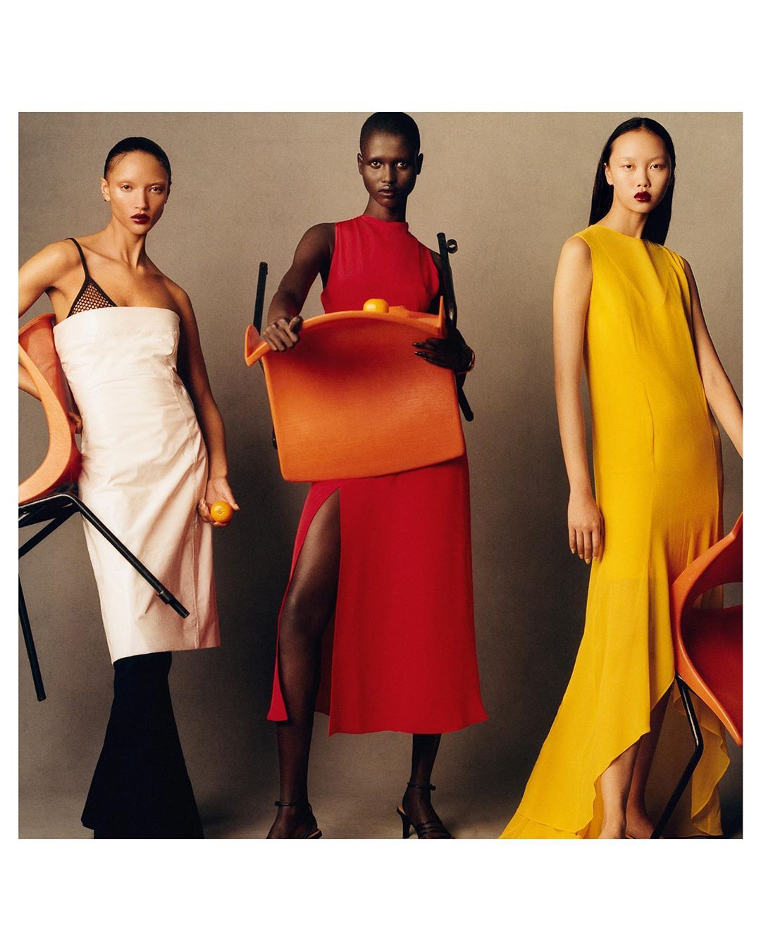 Three models in colorful dresses pose with orange chairs and citrus fruit in a studio setting for a fashion editorial.