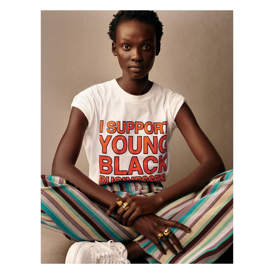 A young Black woman poses in a studio wearing a t-shirt that reads, 'I support young black businesses'. She wears striped pants and sneakers.