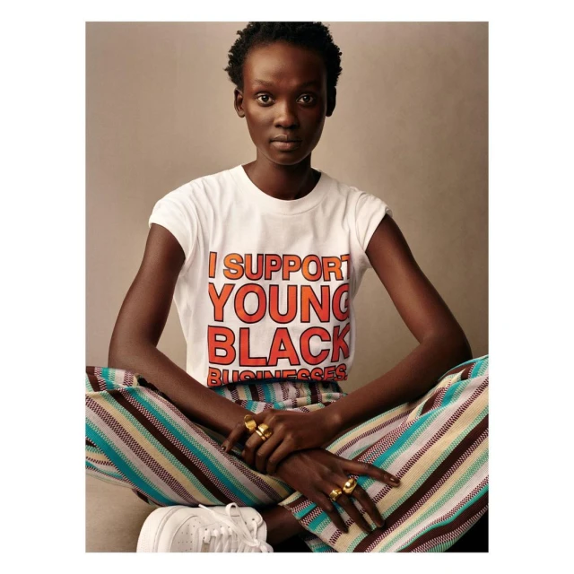 A young Black woman poses in a studio wearing a t-shirt that reads, 'I support young black businesses'. She wears striped pants and sneakers.