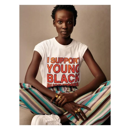 A young Black woman poses in a studio wearing a t-shirt that reads, 'I support young black businesses'. She wears striped pants and sneakers.
