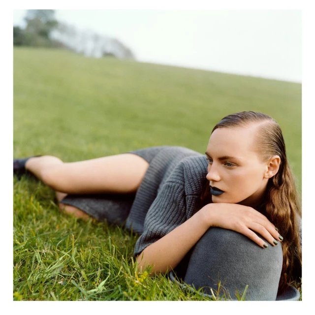 A fair-skinned woman with black lipstick and a gray sweater lying in a grassy field looks off-camera in a beauty and fashion shot.