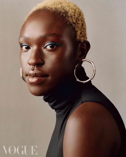 Portrait of a Black woman with short, dyed blonde hair wearing a black turtleneck, silver hoop earrings, and a nose ring on a grey background.