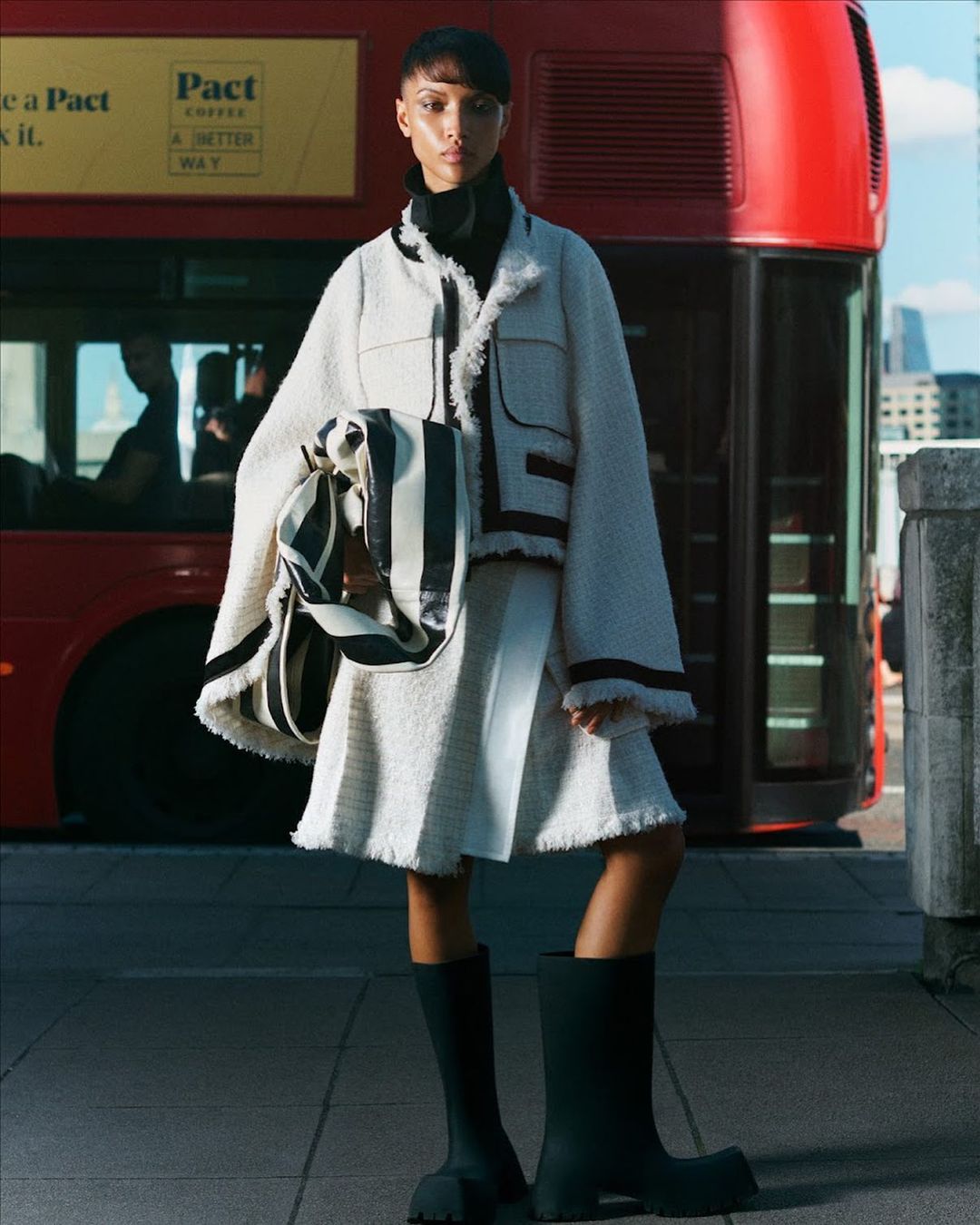 Fashionable woman poses in a tweed jacket and skirt with rubber boots and a striped tote bag in front of a red bus and city.