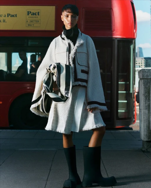 Fashionable woman poses in a tweed jacket and skirt with rubber boots and a striped tote bag in front of a red bus and city.