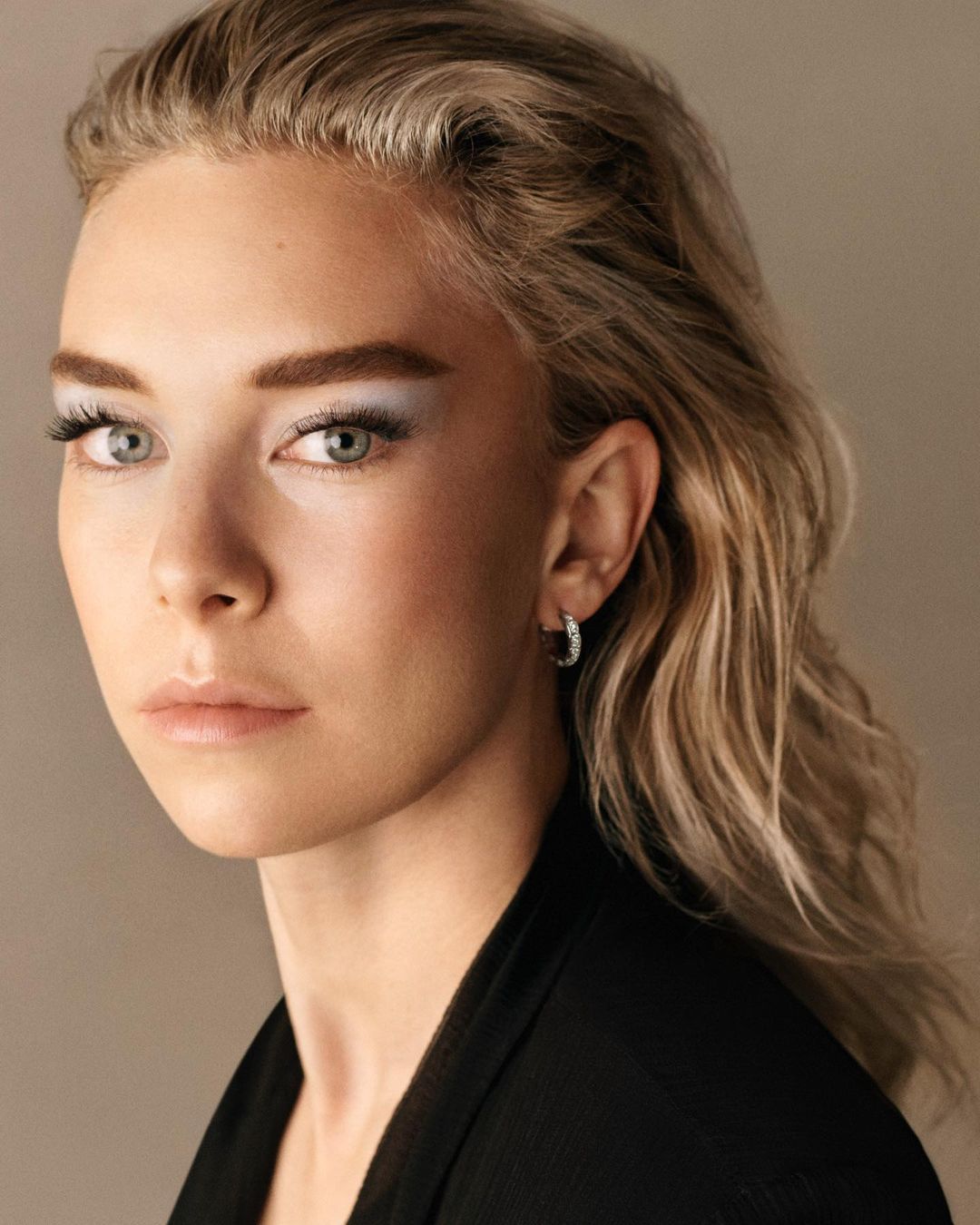 Close-up portrait of a woman with slicked-back blonde hair, pale blue eyeshadow, and hoop earrings against a neutral background.