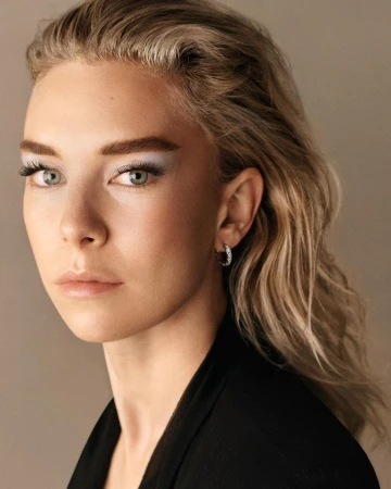 Close-up portrait of a woman with slicked-back blonde hair, pale blue eyeshadow, and hoop earrings against a neutral background.