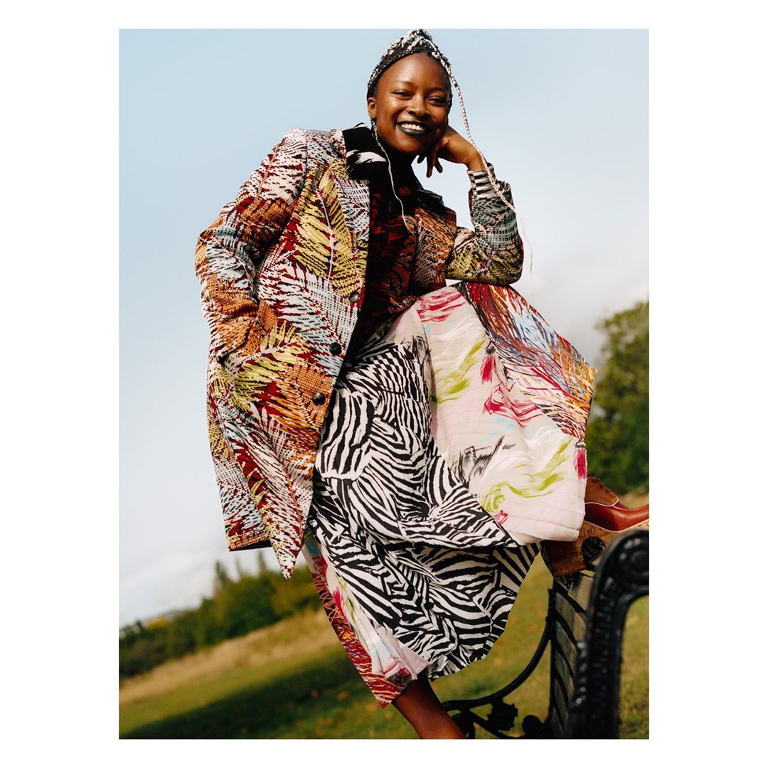 Woman in patterned coat sitting on park bench with a smile, captured in an eye-level shot with a light sky and green trees in background.