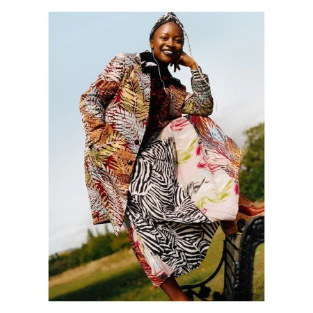 Woman in patterned coat sitting on park bench with a smile, captured in an eye-level shot with a light sky and green trees in background.