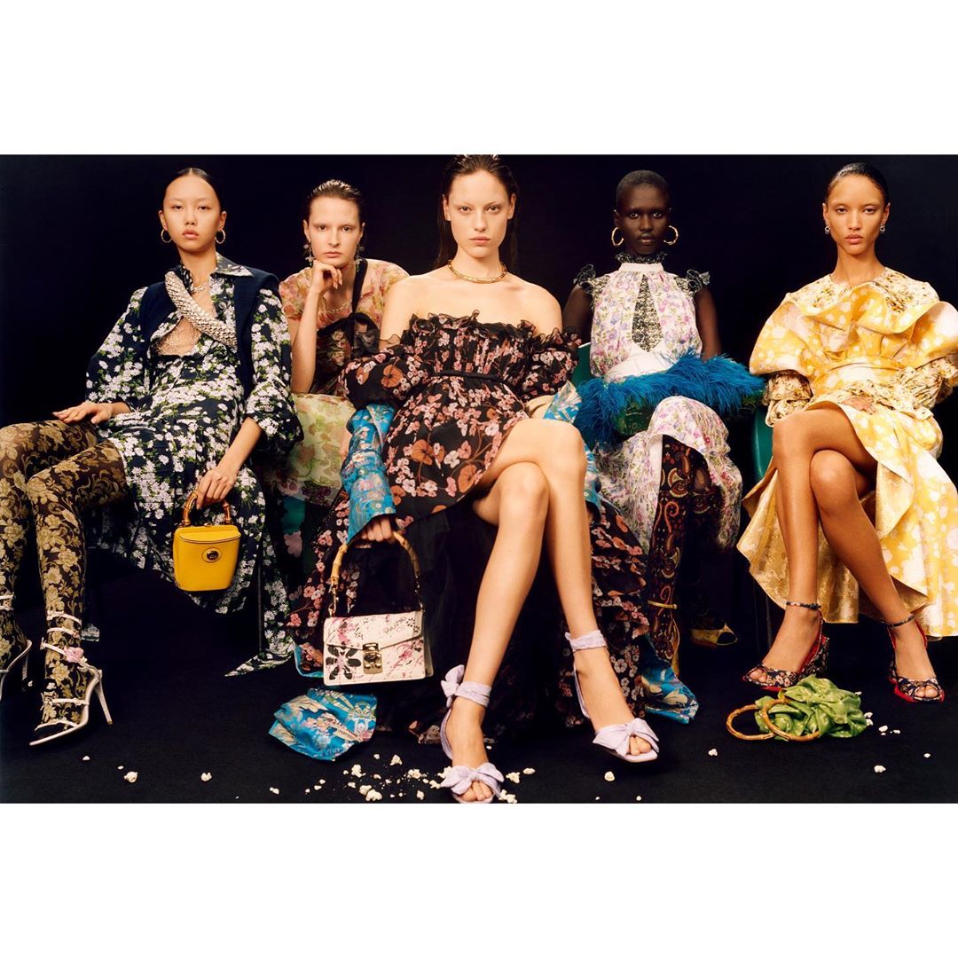 Five female fashion models in floral print dresses and heels are seated in a line against a dark background, holding various handbags.