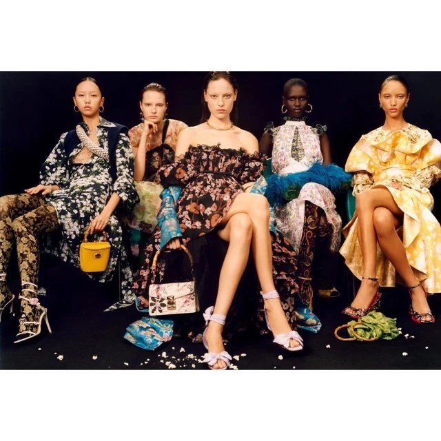 Five female fashion models in floral print dresses and heels are seated in a line against a dark background, holding various handbags.