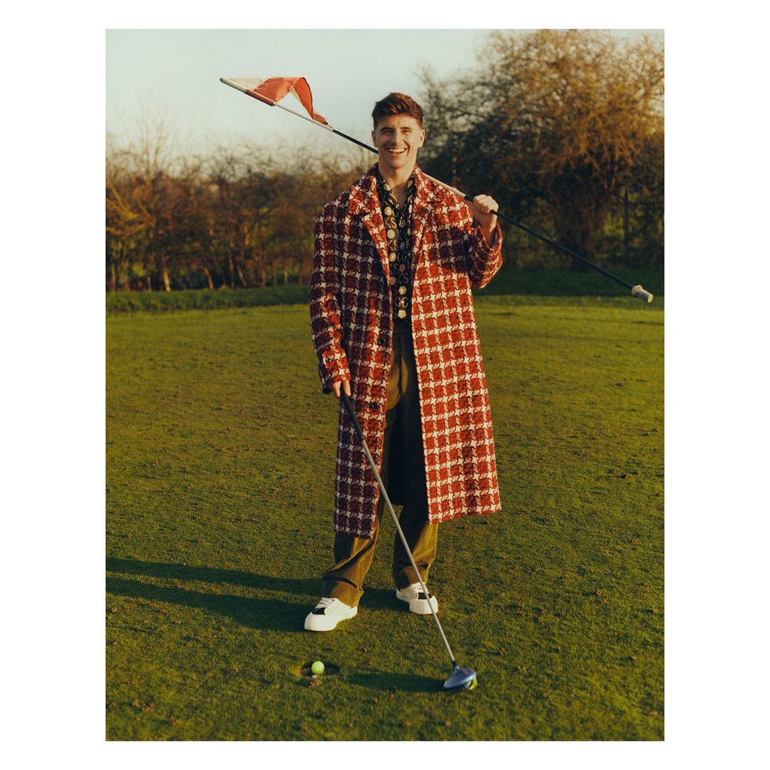 A male golfer poses on a sunny green course in a red-and-white coat, holding a club with the flag over his shoulder in daytime.