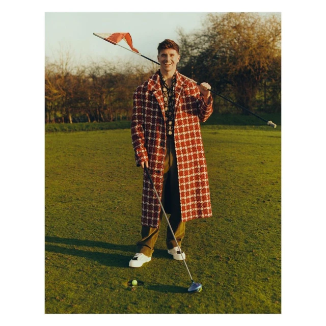 A male golfer poses on a sunny green course in a red-and-white coat, holding a club with the flag over his shoulder in daytime.