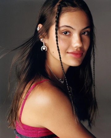 Brunette woman in close-up portrait with braided hair details, wearing jewelry and a sleeveless top against a grey backdrop.