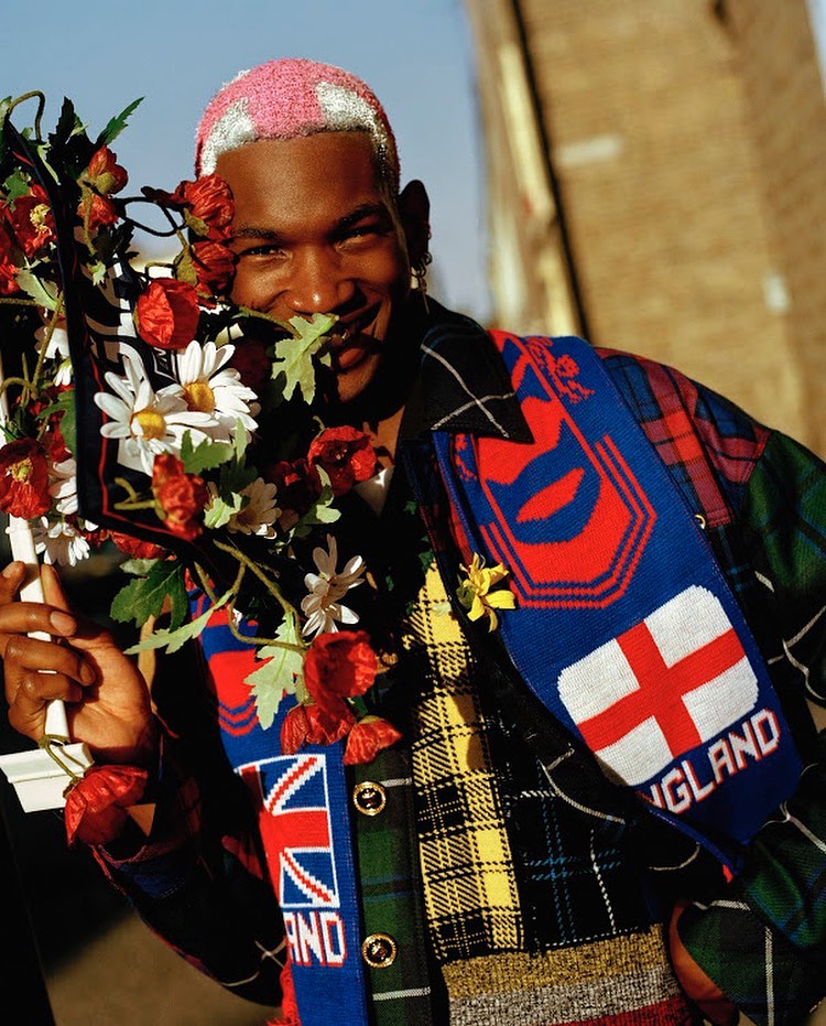 Stylish man in a plaid jacket and pink-tinted hair holds flowers with England-themed scarfs on a sunny day in an urban setting.