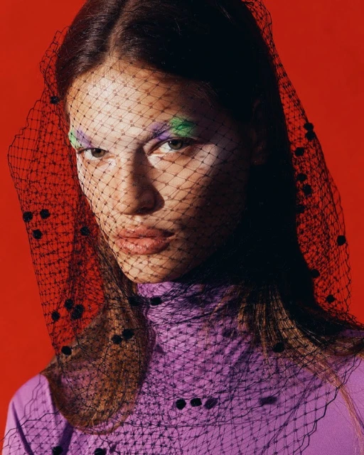 A young woman with bold makeup poses against a red background, her face partially covered by a black polka-dot net veil.