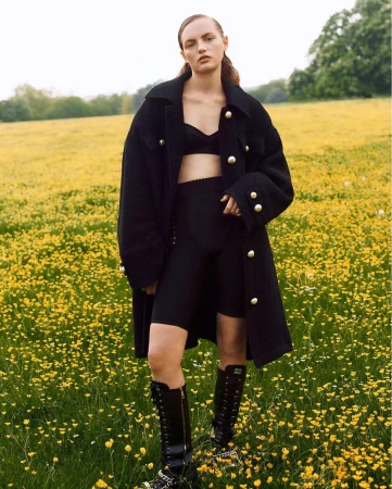 A woman in a black coat, shorts, and bra poses in a field of yellow flowers, showcasing a fashion editorial style in natural light.