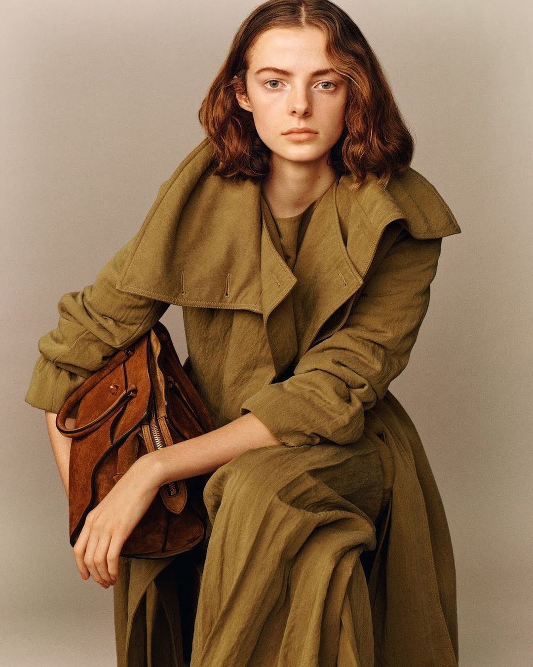 Portrait of a young fair-skinned woman in an olive green coat and dress holding a brown leather bag, in a studio setting.
