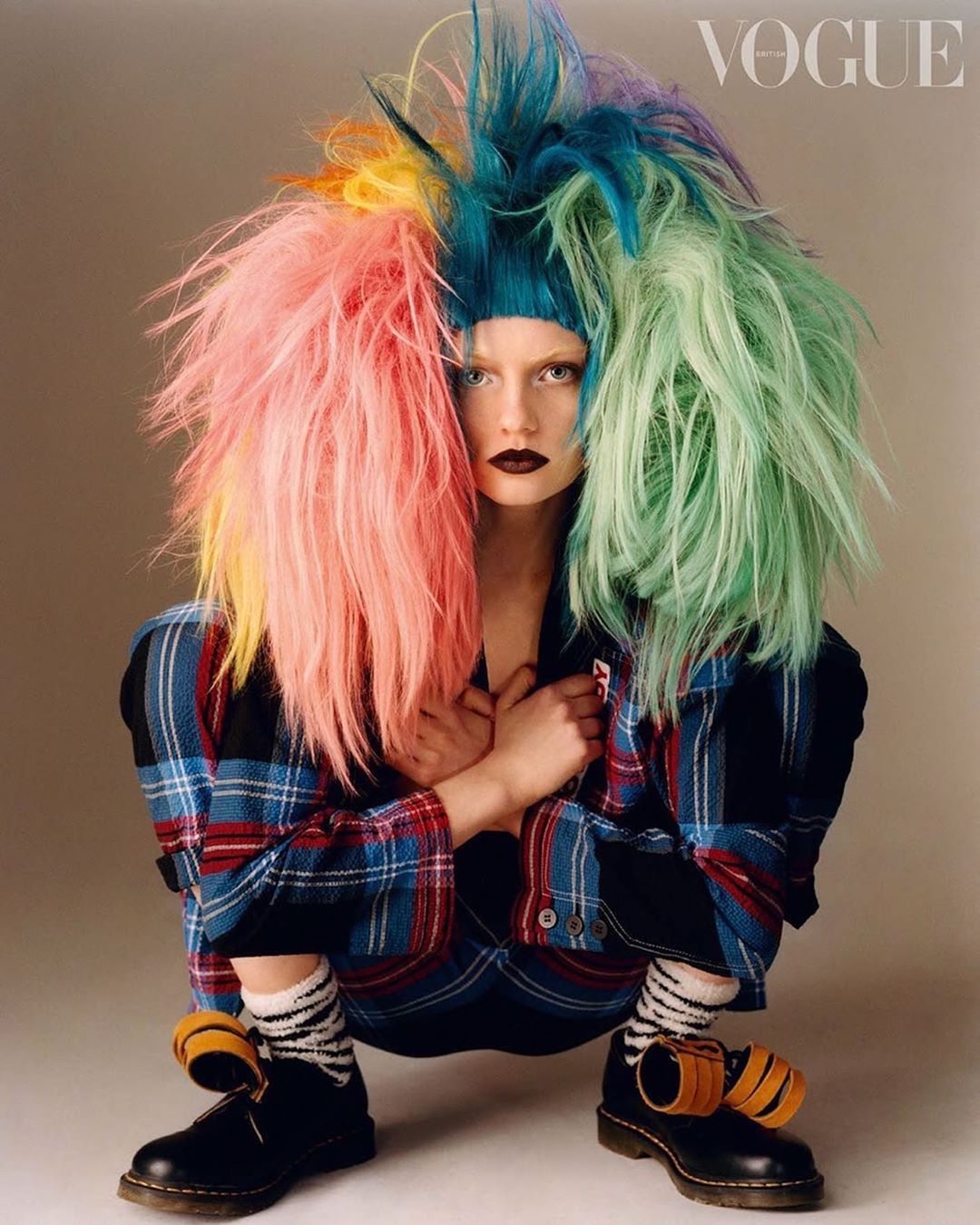 A young adult woman with striking dark lipstick is crouching while posing in a plaid jacket and colorful hair for a fashion editorial.