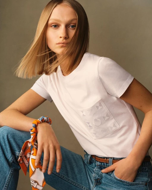A woman with blonde hair poses in a studio wearing a white t-shirt and blue jeans with a scrunchie on her wrist.