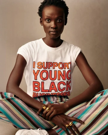Portrait of an African American woman in a statement t-shirt and striped pants sitting in front of a neutral backdrop in a studio setting.