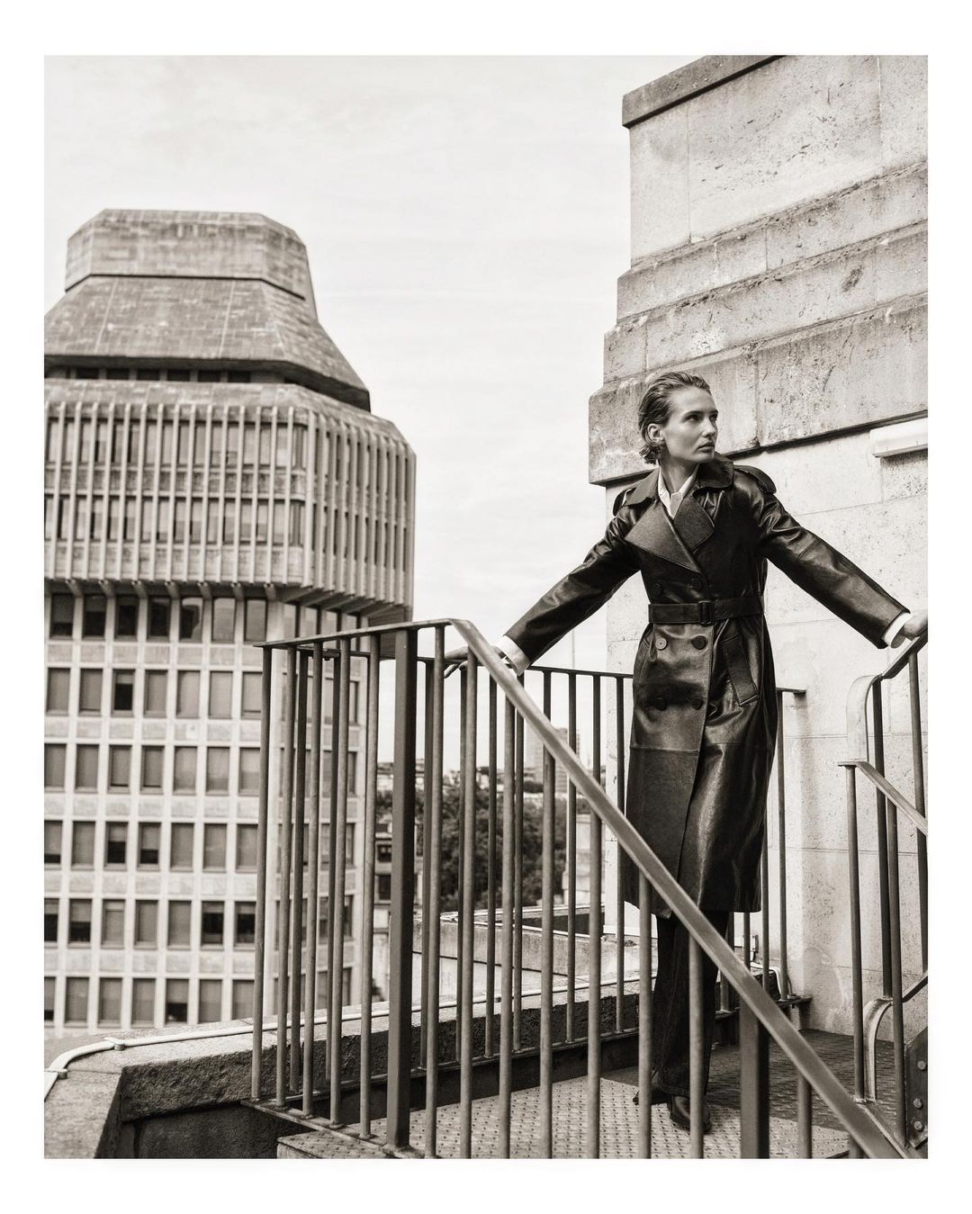 Fashion shot of a woman in a long black coat posing on rooftop stairs with a high-rise building in the background, editorial style.