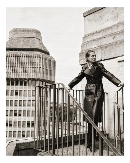 Fashion shot of a woman in a long black coat posing on rooftop stairs with a high-rise building in the background, editorial style.