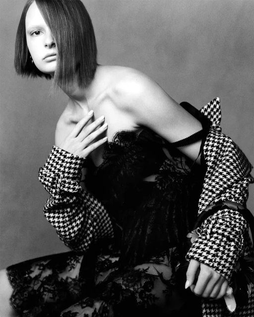 Monochrome fashion portrait of a woman with a bob haircut wearing a lace dress and a houndstooth coat in a studio setting.
