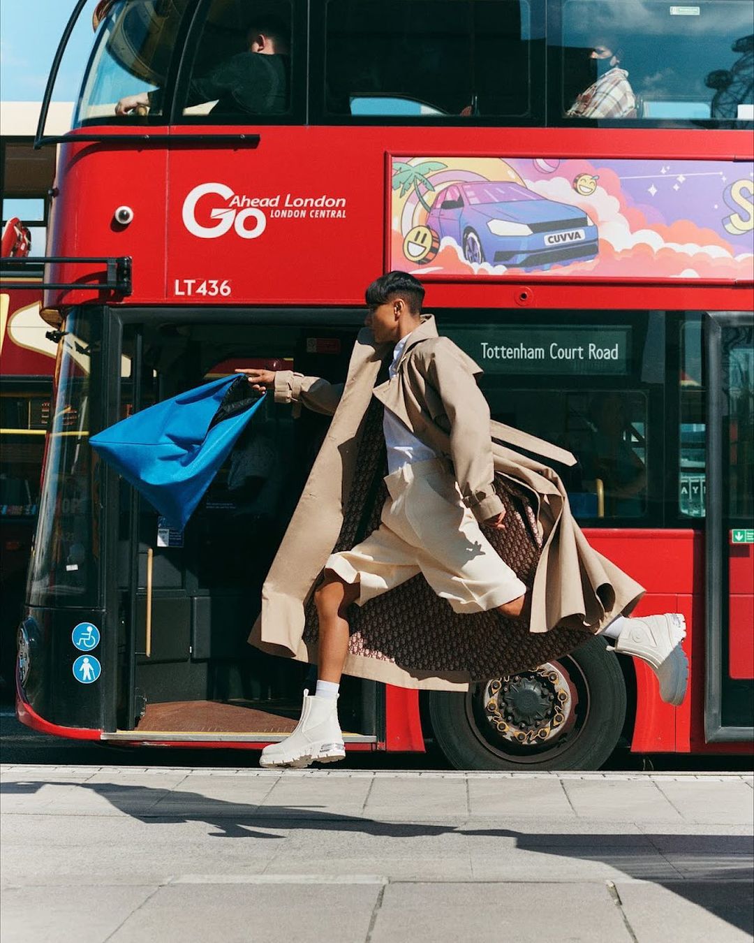 A fashionable adult male runs by a red double-decker bus in London, wearing a trench coat, shorts, and boots, carrying a blue bag.