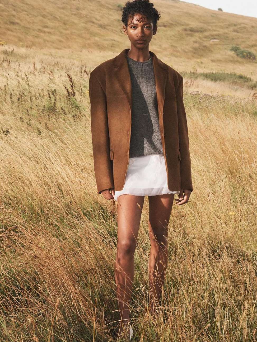 A young adult woman with short curly hair models a brown blazer, gray sweater, and white skirt in a golden field, standing outdoors.