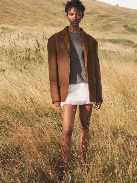 A young adult woman with short curly hair models a brown blazer, gray sweater, and white skirt in a golden field, standing outdoors.