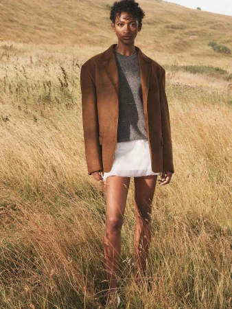 A young adult woman with short curly hair models a brown blazer, gray sweater, and white skirt in a golden field, standing outdoors.