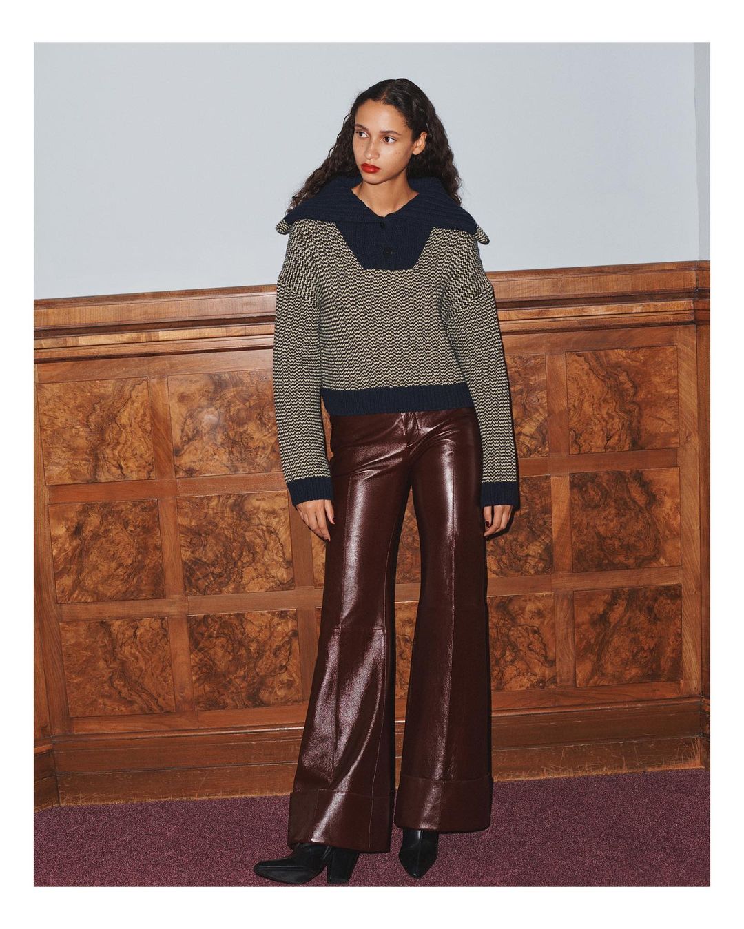 A woman models a patterned knit sweater with a large collar and wide-leg vinyl pants, posing in front of a textured wooden wall and mauve carpet.