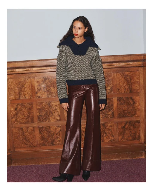 A woman models a patterned knit sweater with a large collar and wide-leg vinyl pants, posing in front of a textured wooden wall and mauve carpet.