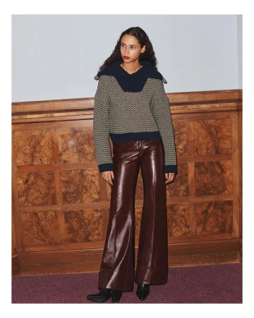 A woman models a patterned knit sweater with a large collar and wide-leg vinyl pants, posing in front of a textured wooden wall and mauve carpet.